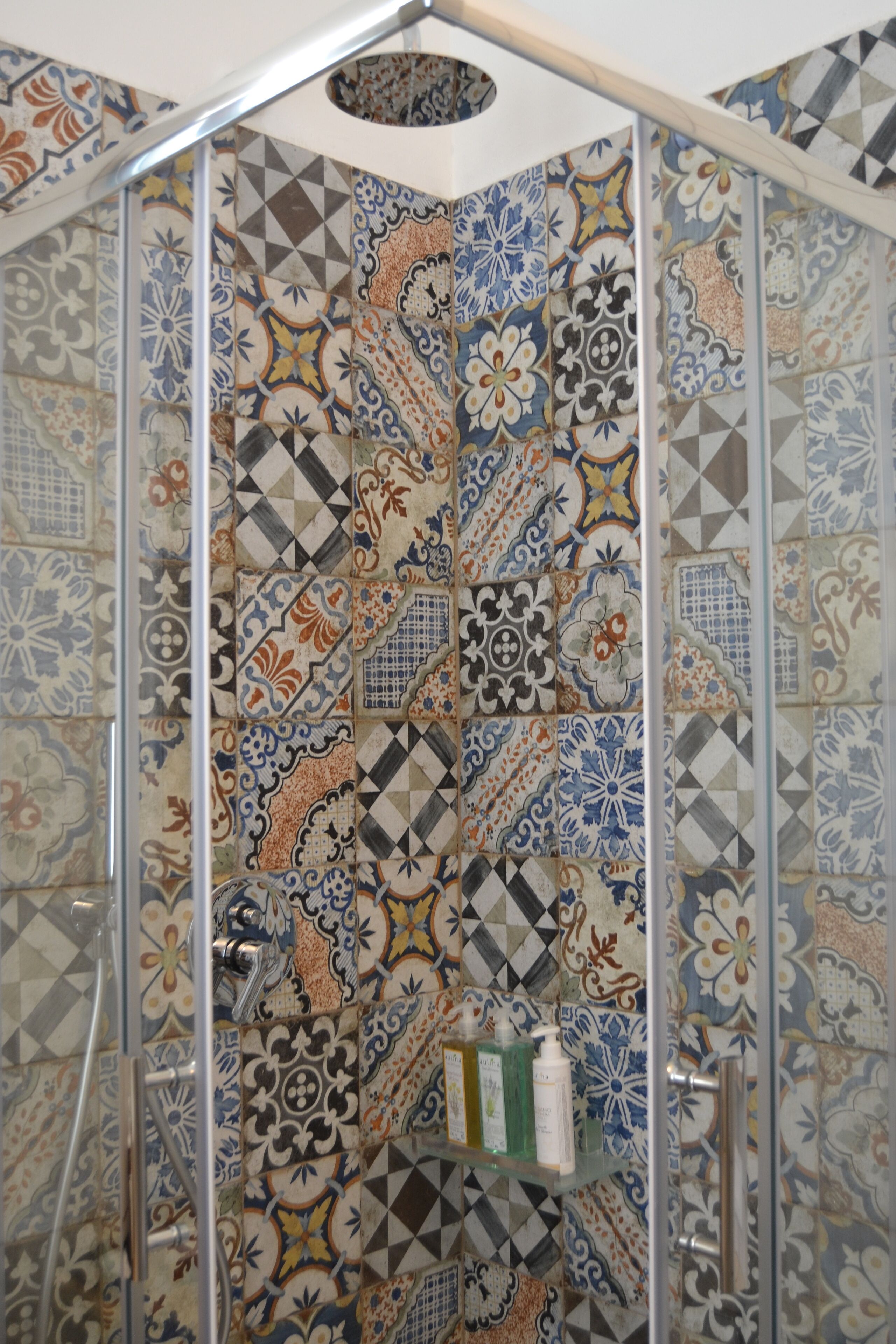 A shower area with a glass door and a tiled wall featuring a variety of geometric and floral patterns.