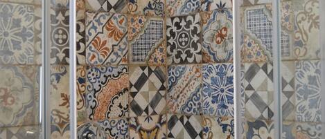 A shower area with a glass door and a tiled wall featuring a variety of geometric and floral patterns.