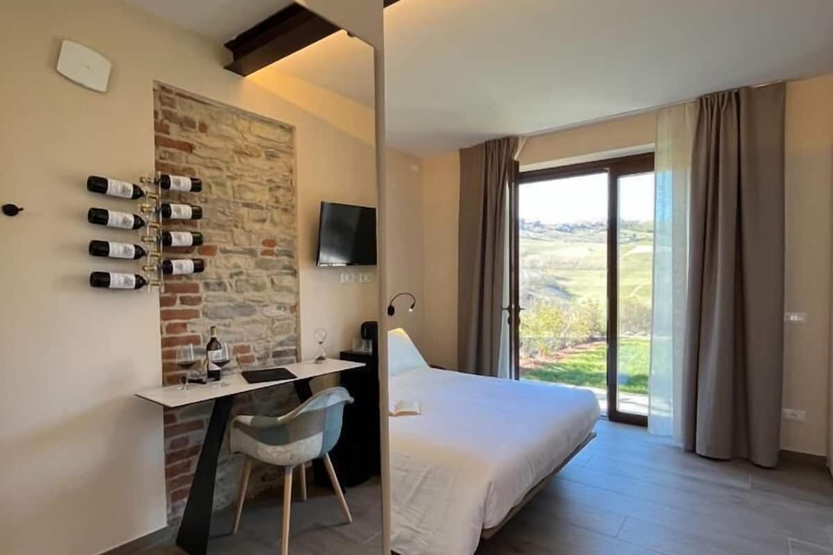 A modern hotel room with a bed, a desk with a chair, a flat-screen TV, a stone accent wall, and a large window with curtains.