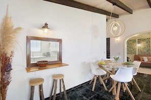 A dining area with a glass table, white chairs, a wooden stool, a mirror, and a hanging light fixture.