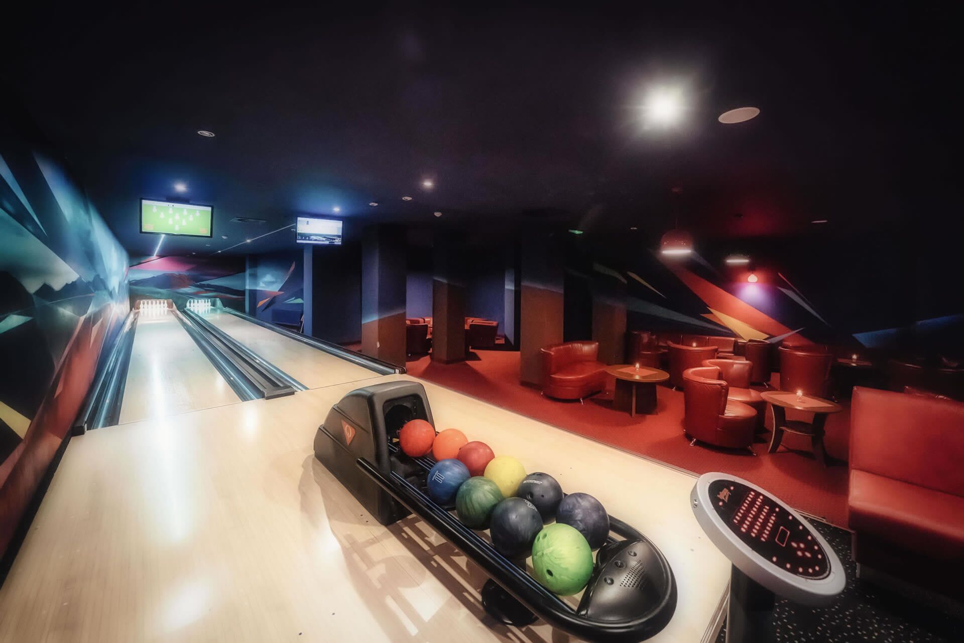 A bowling alley with a row of colorful bowling balls and a bowling pinsetter. There are red seating areas and a TV screen displaying a sports event.