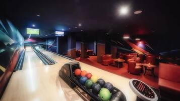 Une salle de bowling avec une rangée de boules colorées et un appareil pour remettre les quilles en place. On y trouve des zones d'assise rouges et un écran de télévision diffusant un événement sportif.