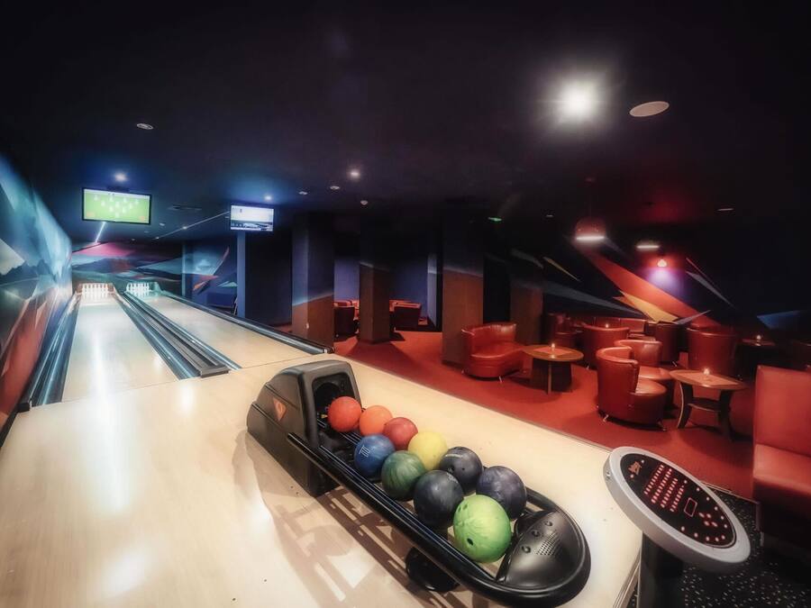 A bowling alley with a row of colorful bowling balls and a bowling pinsetter. There are red seating areas and a TV screen displaying a sports event.