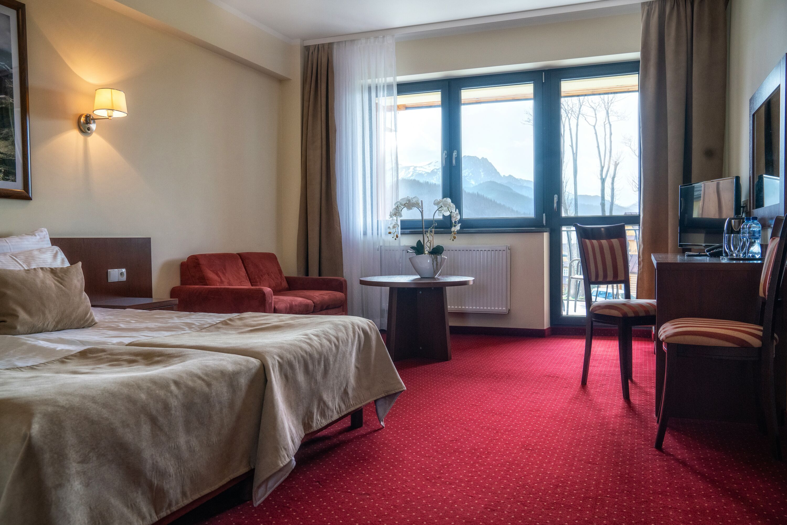 Una habitación de hotel con cama, dos sillones rojos, una mesita con un florero, un escritorio con silla y un ventanal con vistas a la montaña.