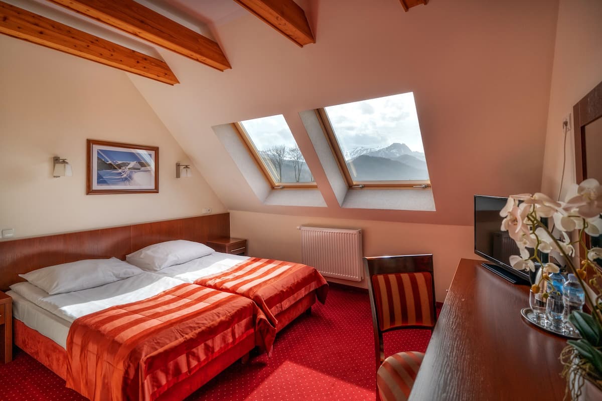A hotel room with two beds, a TV, a desk, a chair, and a view of mountains through skylights.