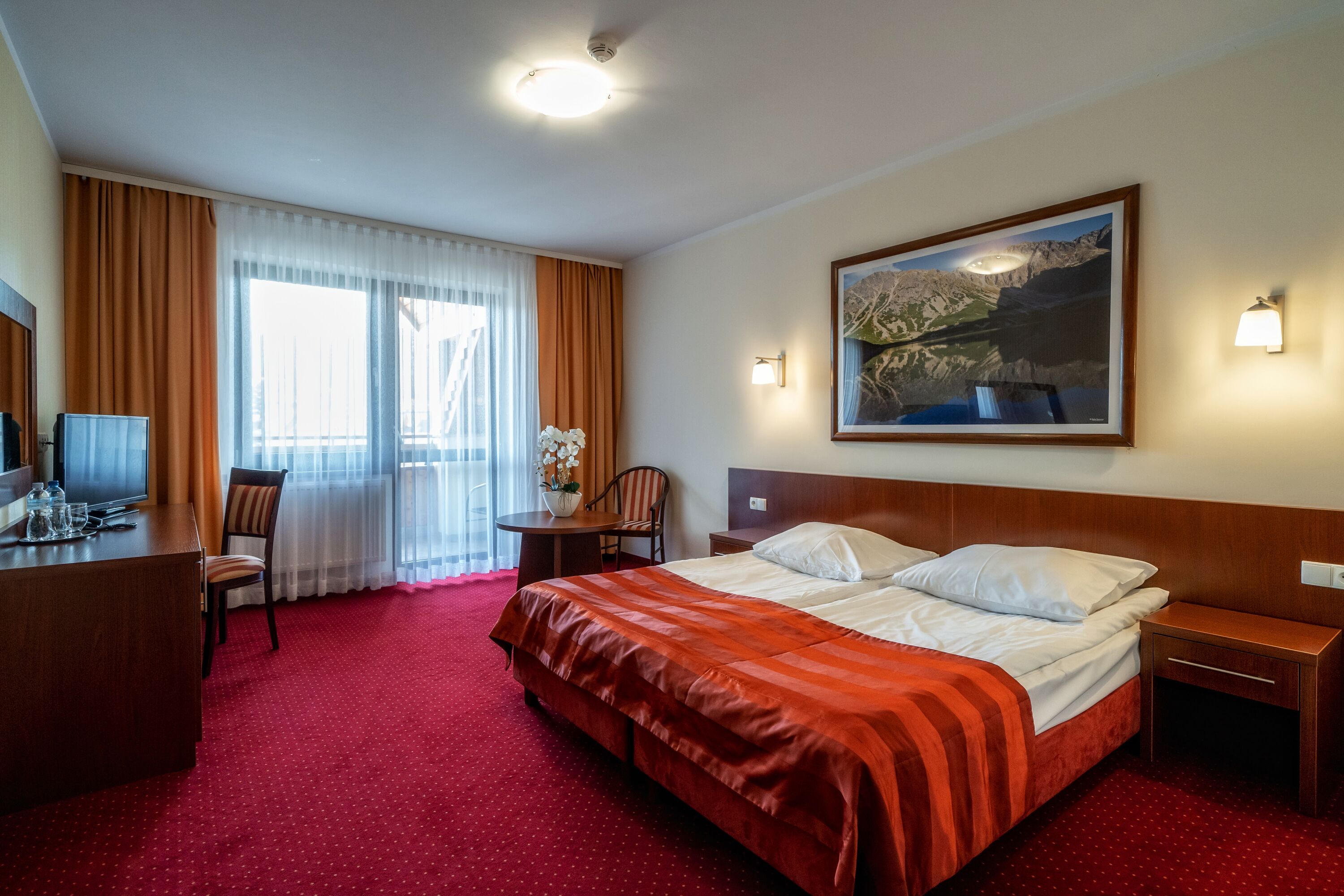 Habitación de hotel con una cama grande, un escritorio, una silla, una mesita con un florero y un cuadro en la pared.