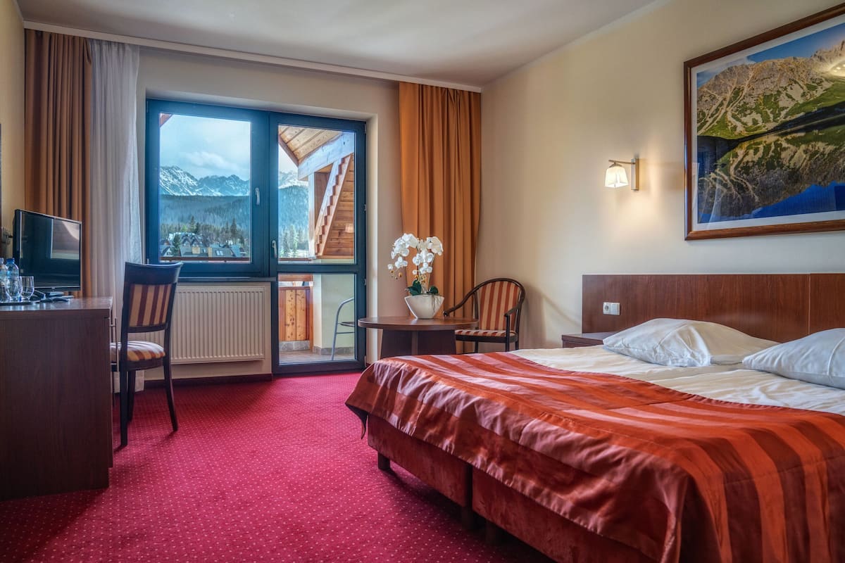 A hotel room with a large bed, a desk, a chair, a small table with a vase of flowers, and a view of mountains through a large window.