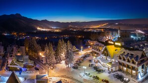 Un hotel cubierto de nieve con una torre destacada, rodeado de árboles cargados de nieve y una vista de un pueblo lejano.