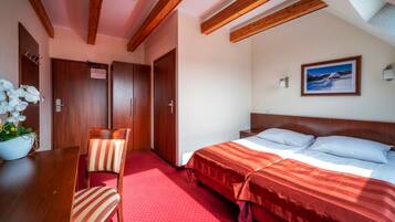 A hotel room with a wooden ceiling, a bed with red bedding, a wooden nightstand, a chair, and a wall-mounted picture.