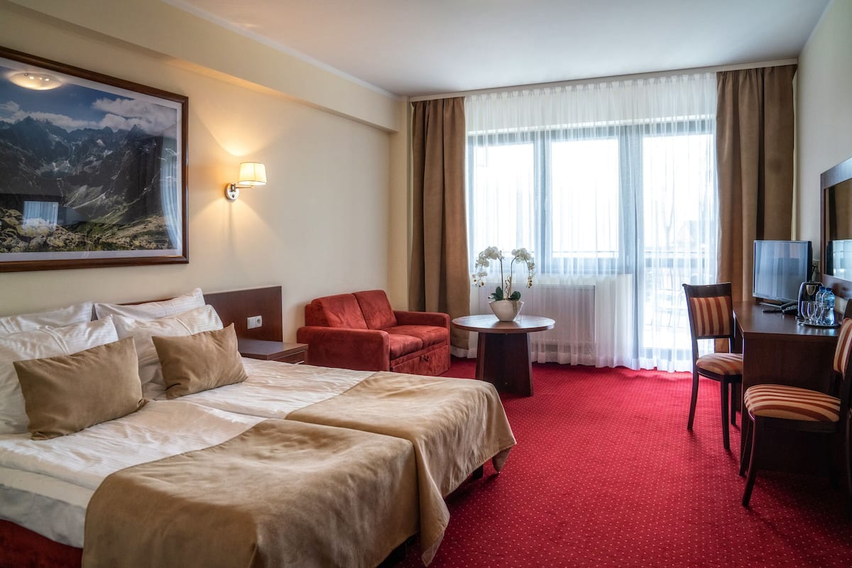A hotel room with a large bed, two bedside tables, a desk with a chair, a small table with a plant, and a large window with curtains.