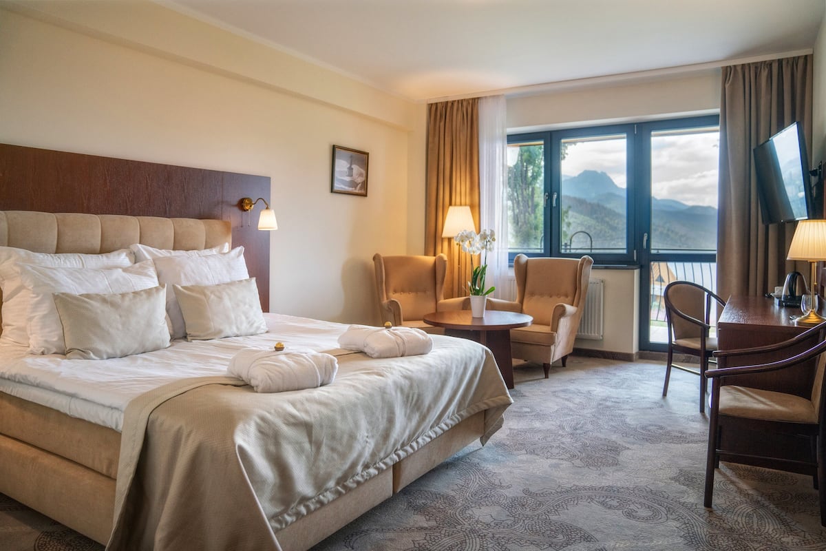 A hotel room with a large bed, a desk, two chairs, a window with a mountain view, and a TV.