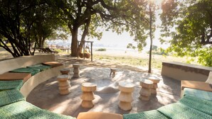 A circular outdoor seating area with green patterned cushions, wooden stools, and a view of a beach and water in the background. - Zikomo Safari Lodge (South Luangwa National Park)