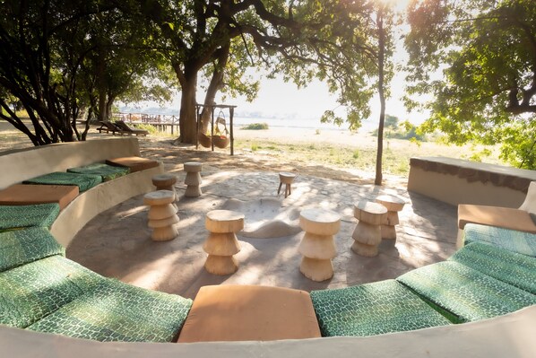 A circular outdoor seating area with green patterned cushions, wooden stools, and a view of a beach and water in the background. - Zikomo Safari Lodge (South Luangwa National Park)