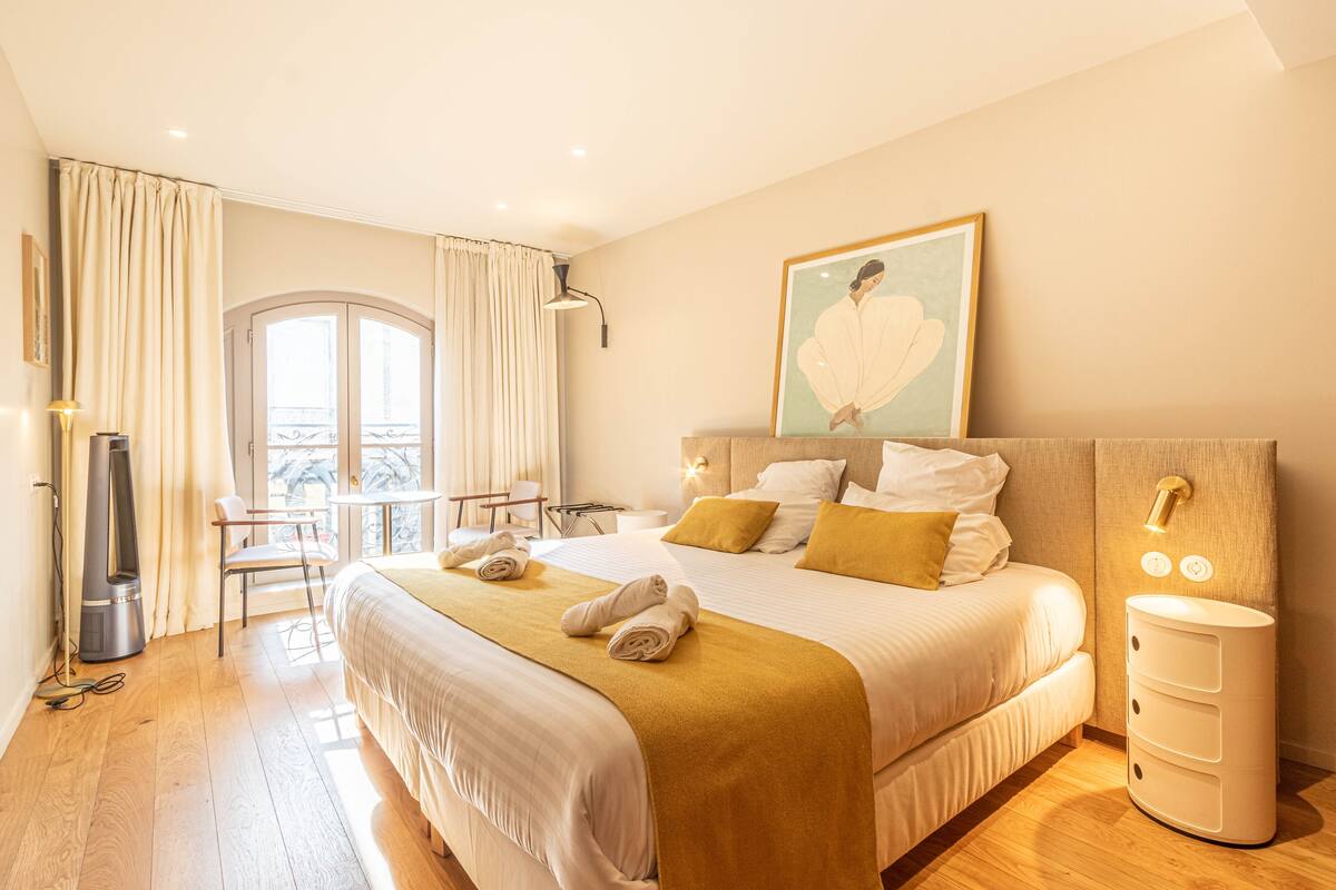 Une chambre d’hôtel avec un grand lit, une table de chevet, une chaise, une fenêtre avec des rideaux et un balcon donnant sur une vue agréable.