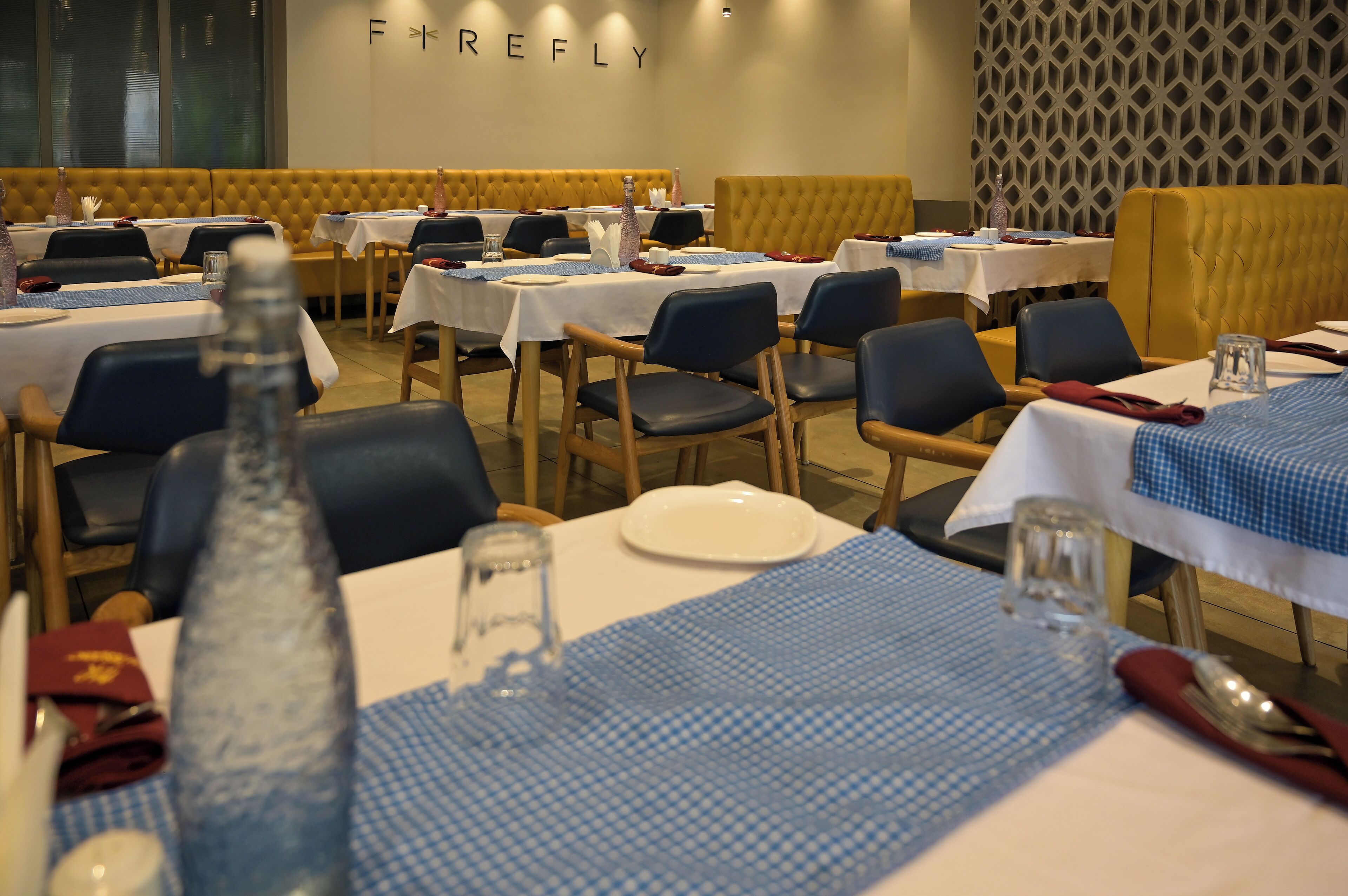 Un restaurant où les tables sont dressées pour le service, avec un mélange de nappes blanches et bleues, et un mur à motifs en arrière-plan.