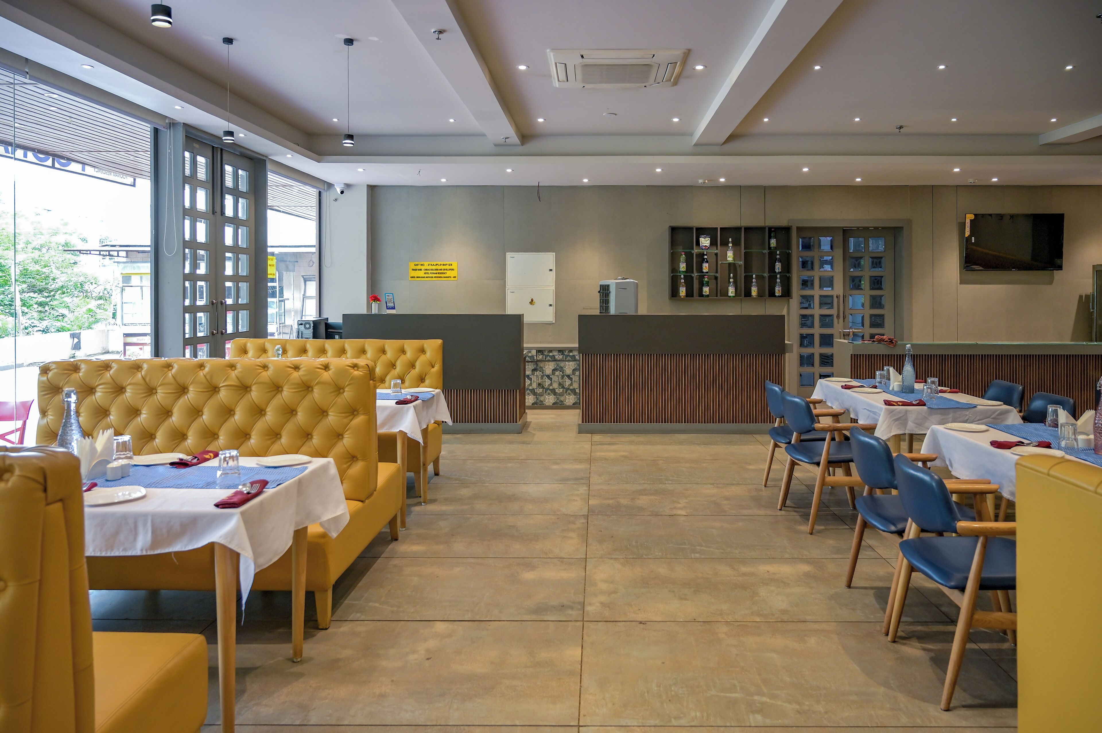 A restaurant with yellow upholstered booths and tables, a large window, and a television on the wall.