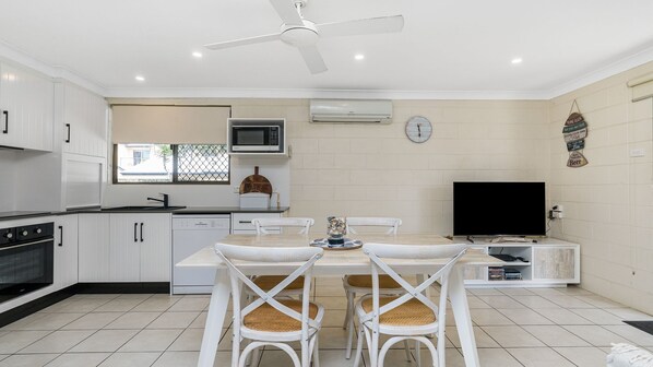 A modern kitchen with white cabinetry, a dining area with a table and chairs, a ceiling fan, and a flat-screen TV.