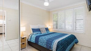 A bedroom with a bed, bedside tables, a window with blinds, and a white brick wall. - Iluka Villa 10 right on the Clarence River (ILUKA)