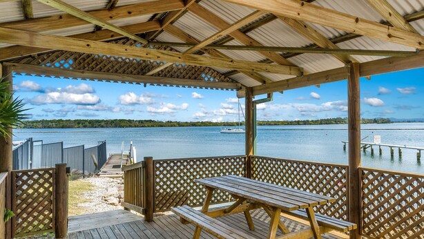 A covered wooden deck with a picnic table, overlooking a calm lake with a sailboat. - Iluka Villa 10 right on the Clarence River (ILUKA)