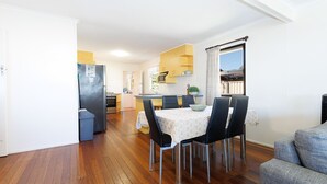 A modern kitchen with wooden cabinets, a dining table with chairs, and a view of the outdoors through a window. - Serenity Now Great Location (Iluka)