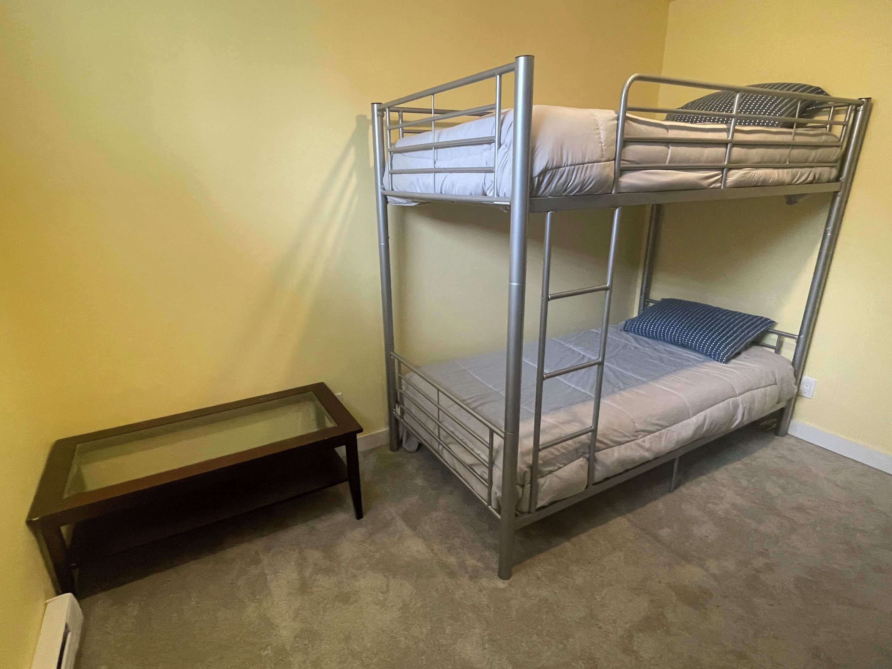 Un letto a castello con un piccolo tavolino accanto, in una stanza dalle pareti spoglie e con pavimento rivestito di moquette.