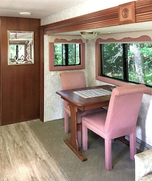 Ein gemütliches Interieur mit einem Holzes Esstisch und rosa gepolsterten Stühlen, ein Fenster mit Blick auf Bäume und ein dekorativer Spiegel an der Wand.