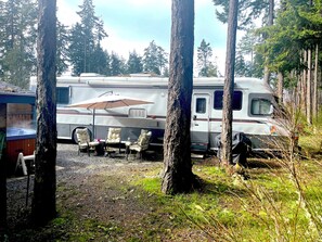 A recreational vehicle (RV) parked in a forested area with a hot tub, outdoor seating, and an umbrella.