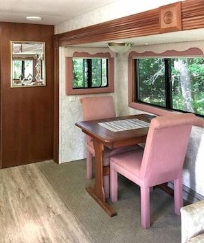 Een knus interieur met een houten tafel en roze gestoffeerde stoelen, een raam met uitzicht op bomen, en een decoratieve spiegel aan de muur.
