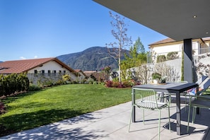 A patio with a table and chairs, a well-maintained lawn, and a mountain view in the background. - Mali Apartment Garden Suite (Tirolo)
