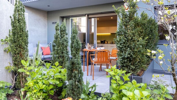 A modern outdoor patio with a dining area, surrounded by various plants and trees. - Mali Appartments Lounge (Tirolo)