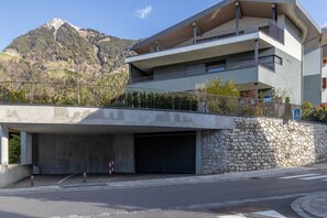 A modern multi-story building with a flat roof, a covered parking area, and a stone wall. - Mali Appartments Lounge (Tirolo)
