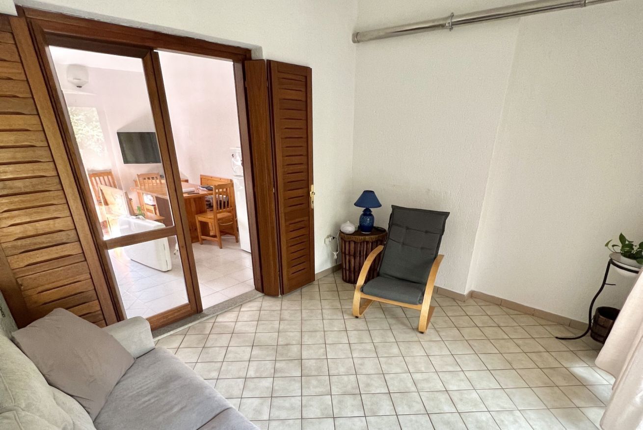 A room with a sliding glass door leading to a balcony, a grey sofa, a wooden chair, and a small table with a lamp.