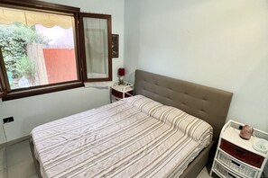 Een slaapkamer met een bed, een nachtkastje, een lamp en een raam met uitzicht op een tuin.
