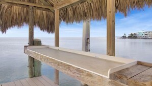 A wooden deck with a thatched roof overlooking calm waters, with a distant view of a resort area.
