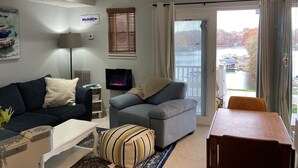A living room with a sofa, a TV, a table, and a view of a lake through a sliding door. - Cozy WATERFRONT condo in a quiet cove.   Private Boat launch (Moneta)