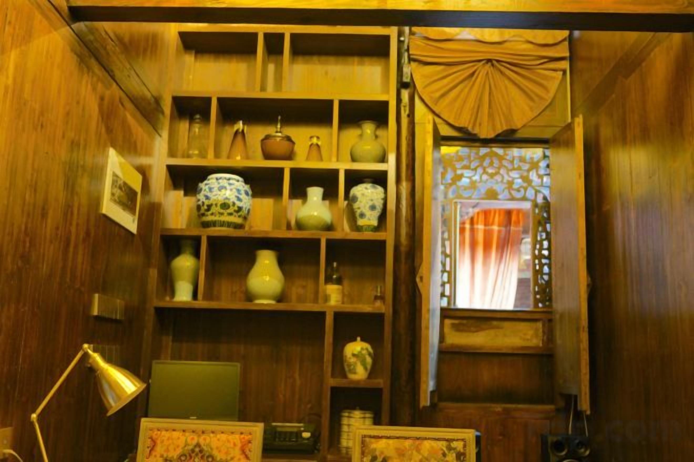 A room with wooden walls, shelves with decorative items, a desk with a lamp, and a window with curtains.