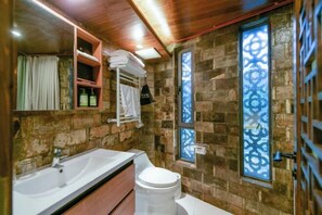 A bathroom with a wooden ceiling, stone walls, a white sink, a toilet, and a window with a decorative pattern. - Ruiqi Xiangyun Inn (Yi County)