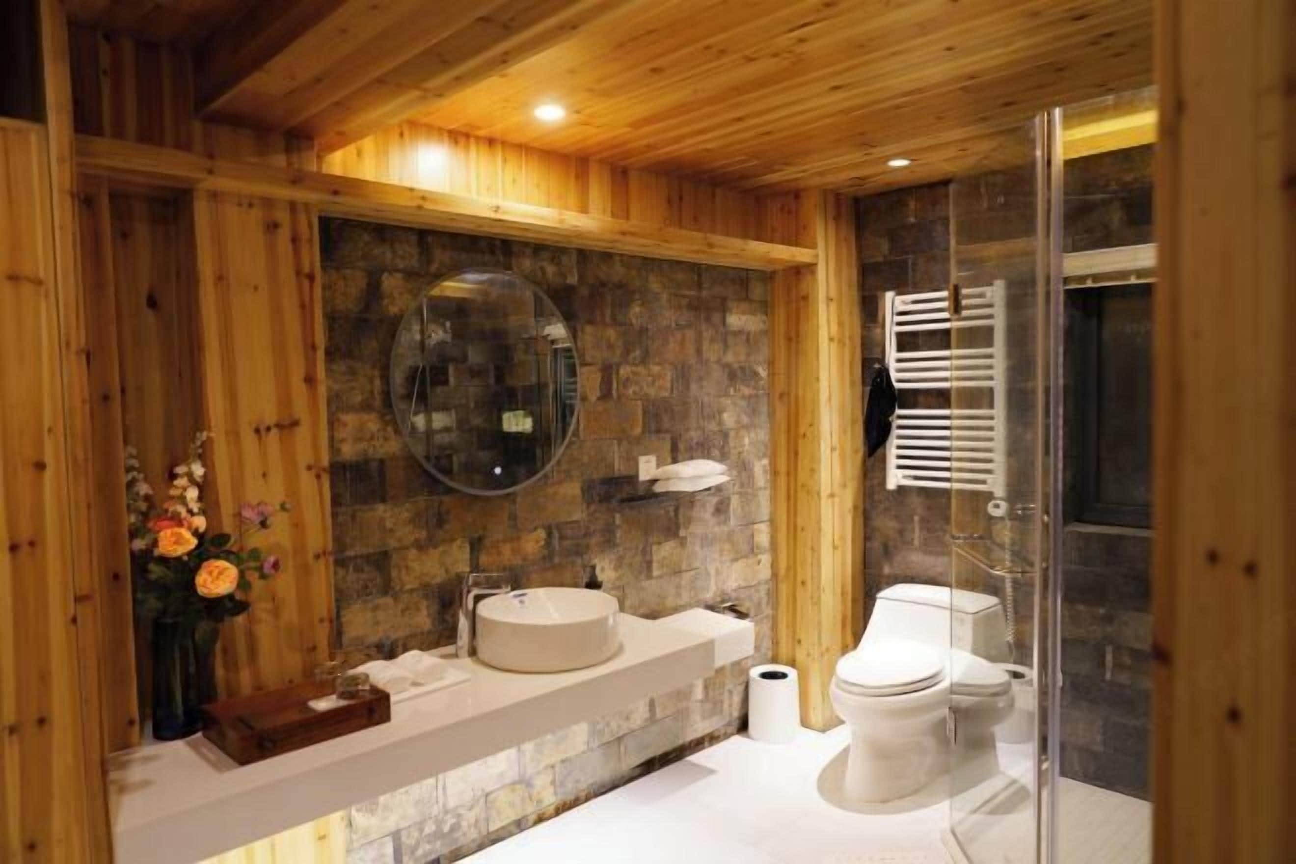 Un bagno con parete in pietra, soffitto in legno, uno specchio rotondo, un lavandino, un WC e un box doccia in vetro.