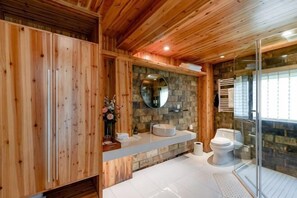 A wooden cabin-style bathroom with a stone wall, a round mirror, a toilet, and a shower area. - Ruiqi Xiangyun Inn (Yi County)