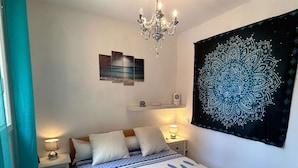 A bedroom with a bed, bedside lamps, a chandelier, a wall tapestry, and a shelf with decorative items.