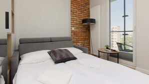 A modern hotel room with a grey upholstered bed, a brick accent wall, a large window with a view of buildings, and a small table with a lamp.