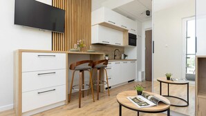 A modern kitchen with white cabinets, wooden accents, and a built-in TV. There are two bar stools and a small round table with a magazine.