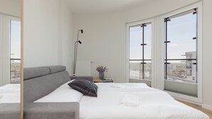 Moderni hotellihuone, jossa on suuri sänky, harmaa sohva, lattiavalaisin ja suuret ikkunat, joista on näkymä kaupunkiin.