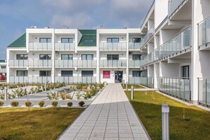 A modern multi-story building with a central entrance, a covered walkway, and a landscaped area with small plants.