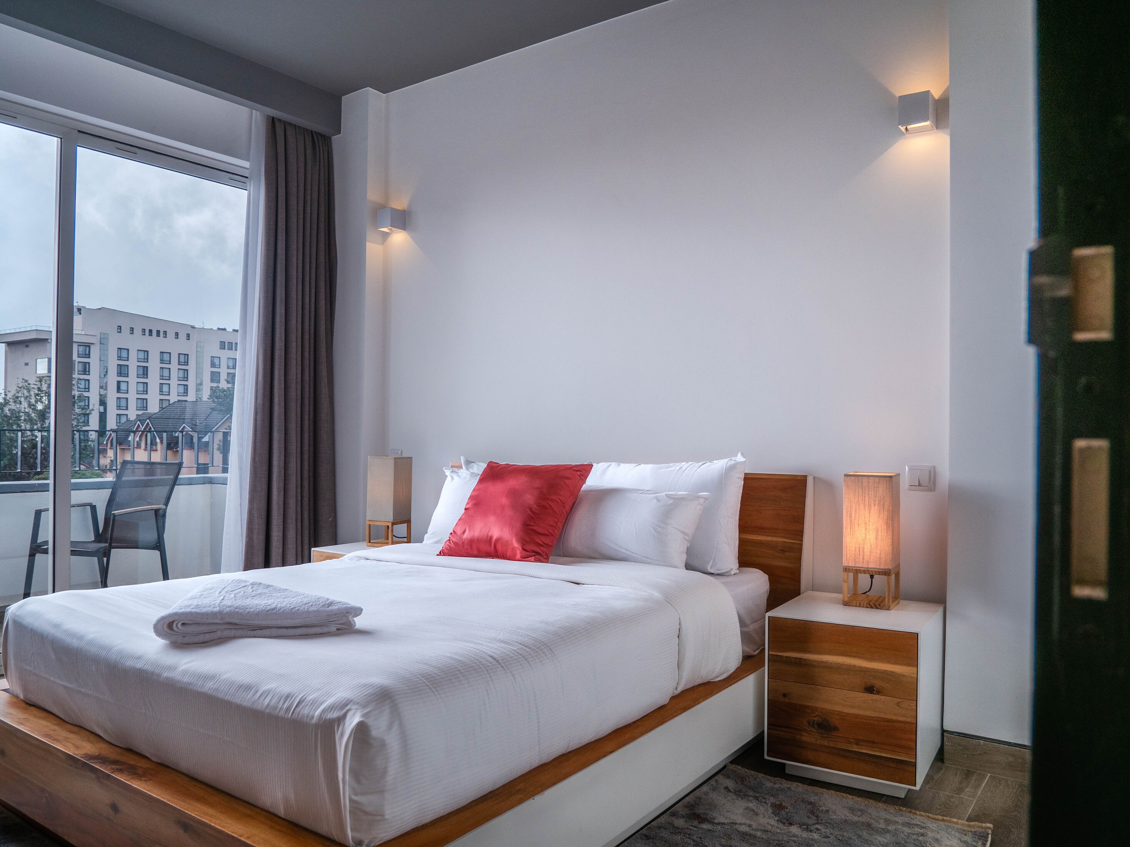 A modern hotel room with a large bed, wooden headboard, bedside tables with lamps, and a view of buildings through a large window.