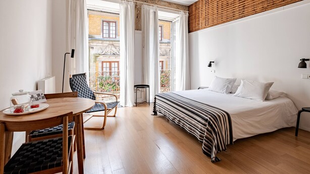 A hotel room with a bed, a dining table with chairs, a small table with a lamp, and a view of the outside balcony. - Artes, the Studio in Porto World Heritage (Porto)