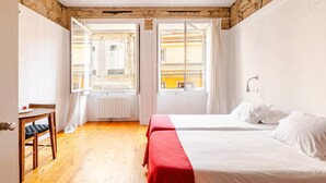 Un dormitorio con piso de madera, una cama con ropa blanca, una colcha roja y una ventana con cortinas.