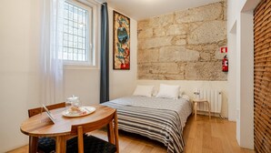 Un dormitorio con pared de piedra, una cama, una mesita de noche, una silla y una ventana con cortinas.