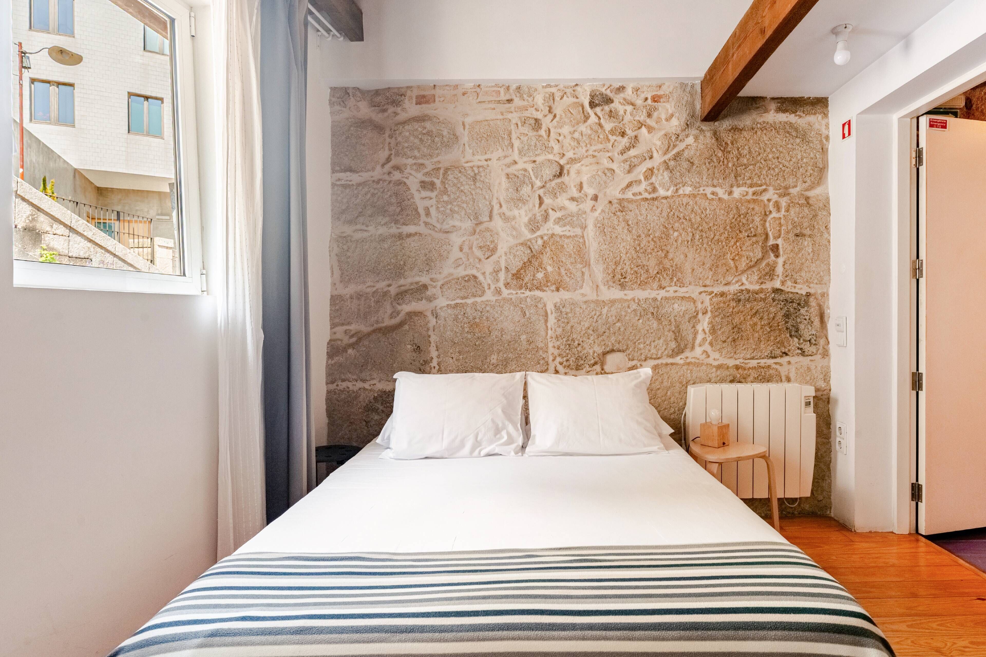 A bedroom with a stone wall, a bed with white bedding, a small table, and a radiator.