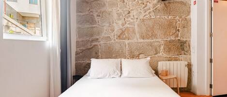 A bedroom with a stone wall, a bed with white bedding, a small table, and a radiator.